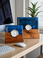 Two square desk clocks with a wood and blue resin design. Each clock has a gold-colored bezel and a white clock face. One clock has the text "Merry Christmas from Tom" engraved on the wood.