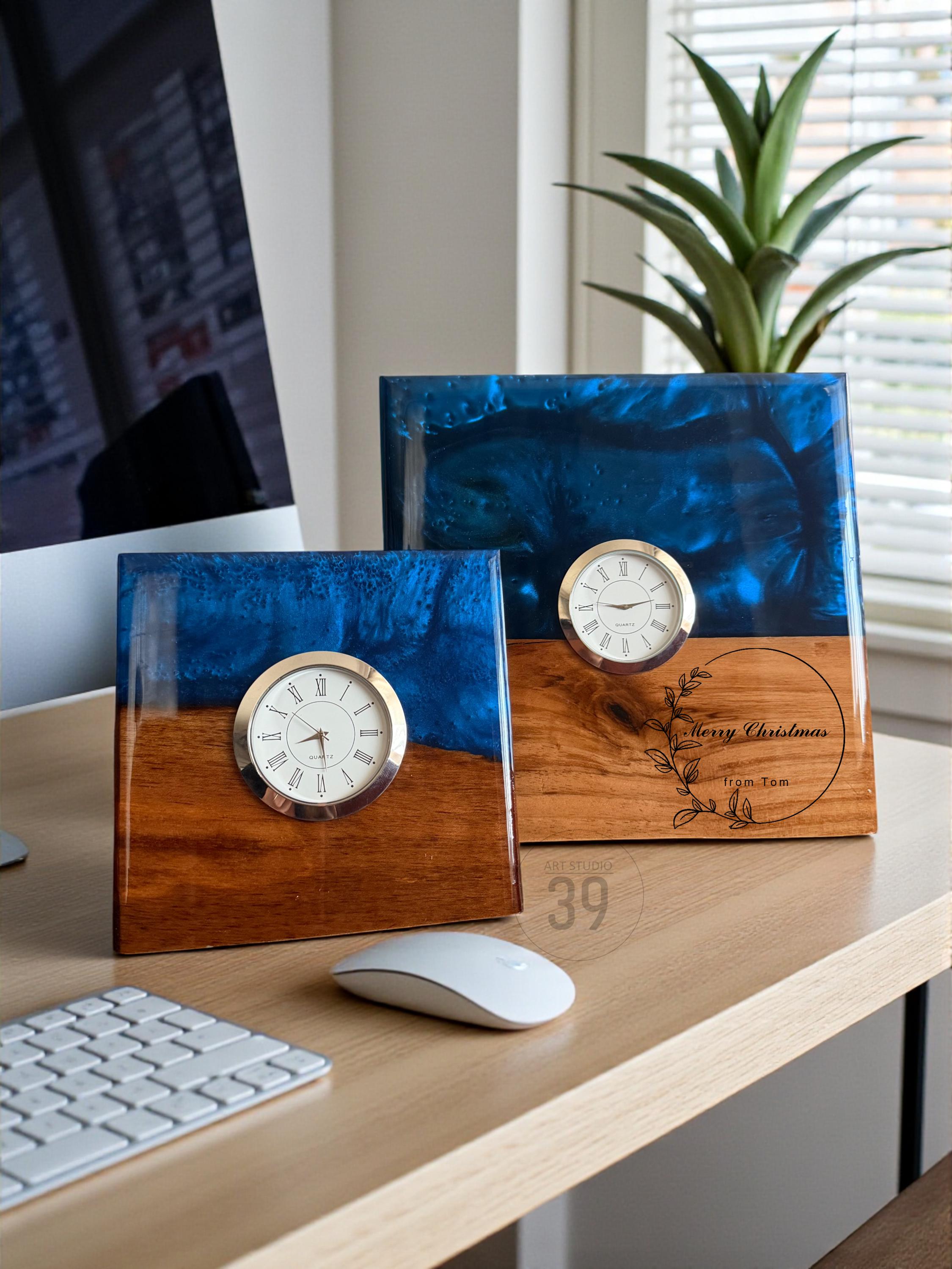 Two square desk clocks with a wood and blue resin design. Each clock has a gold-colored bezel and a white clock face. One clock has the text "Merry Christmas from Tom" engraved on the wood.