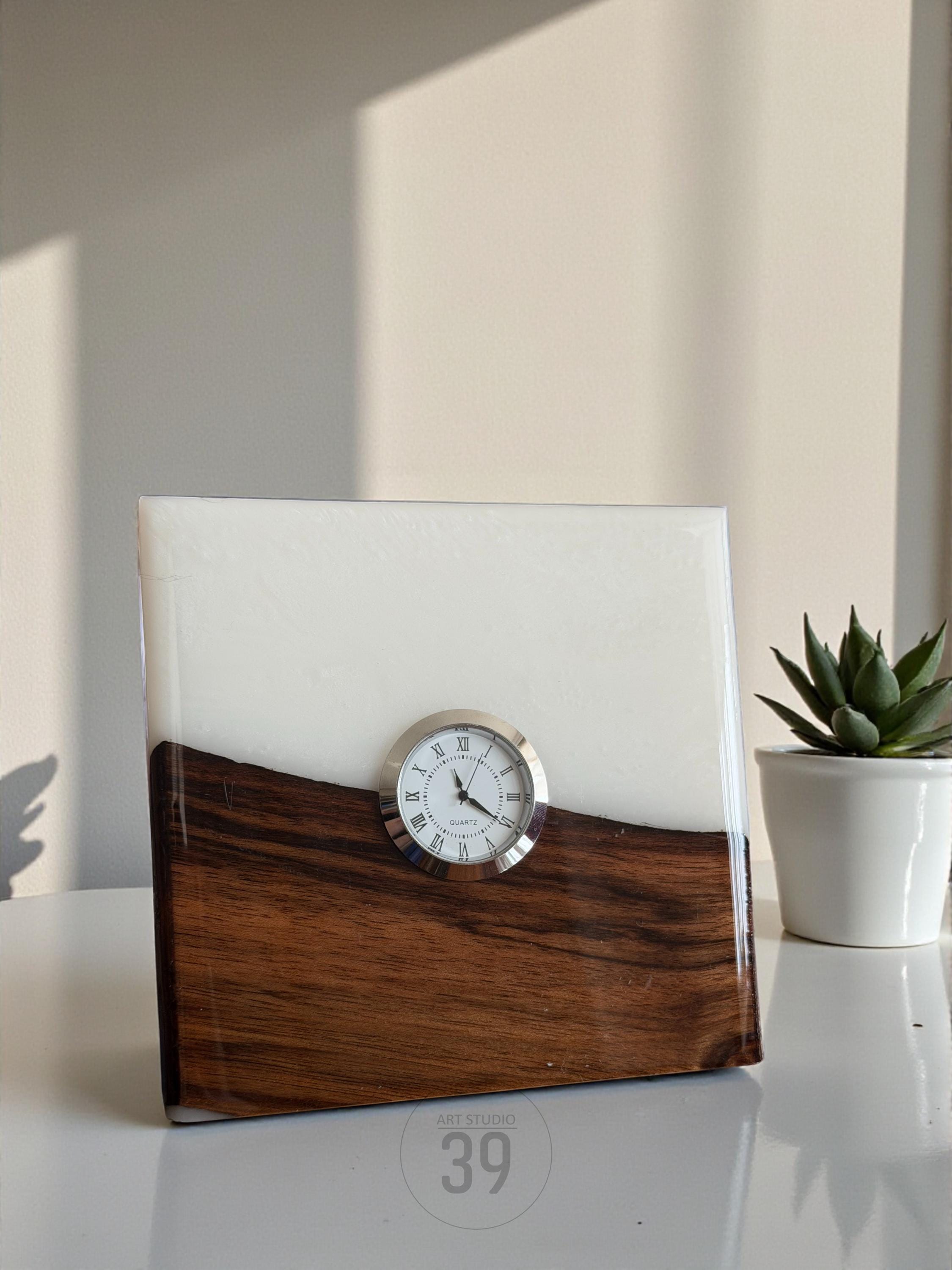 A square clock with a white face and Roman numerals, set in a wooden and white resin block. The clock has a silver bezel. The bottom half is a rich brown wood grain. The text "ART STUDIO 39" is visible.