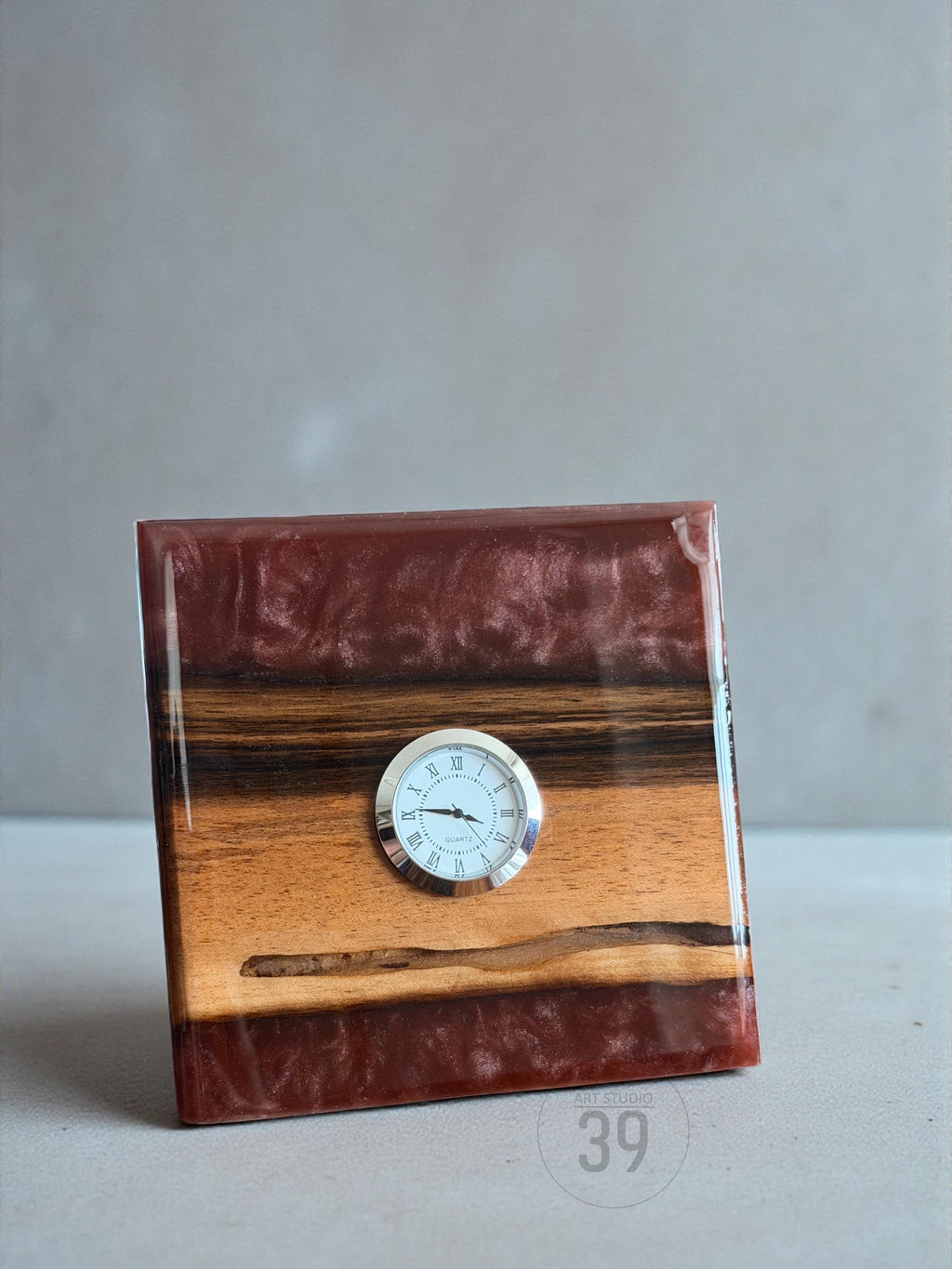 A square wooden clock with a small, round clock face. The clock is made of wood and a reddish-brown resin. The clock face has Roman numerals and black hands. The clock is a decorative item.