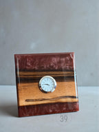 A square wooden clock with a small, round clock face. The clock is made of wood and a reddish-brown resin. The clock face has Roman numerals and black hands. The clock is a decorative item.