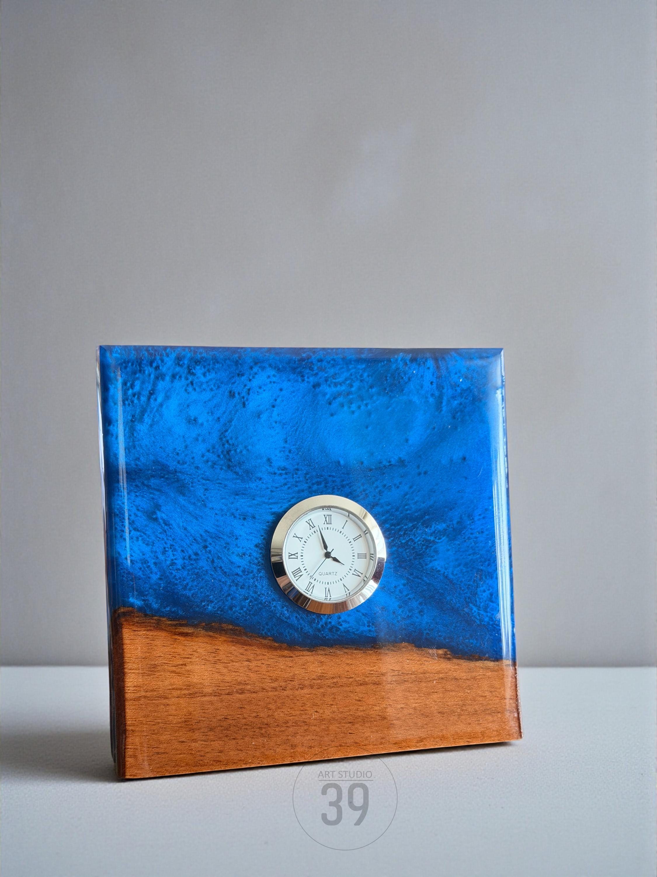 A square clock with a wooden base and a blue resin top. The clock face is white with black Roman numerals and gold-colored hands. The clock is a decorative item.