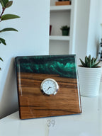 A square wooden clock with a silver-colored clock face. The top portion is a dark green, swirled resin. The bottom is a rich, dark wood. The clock is on a white surface.