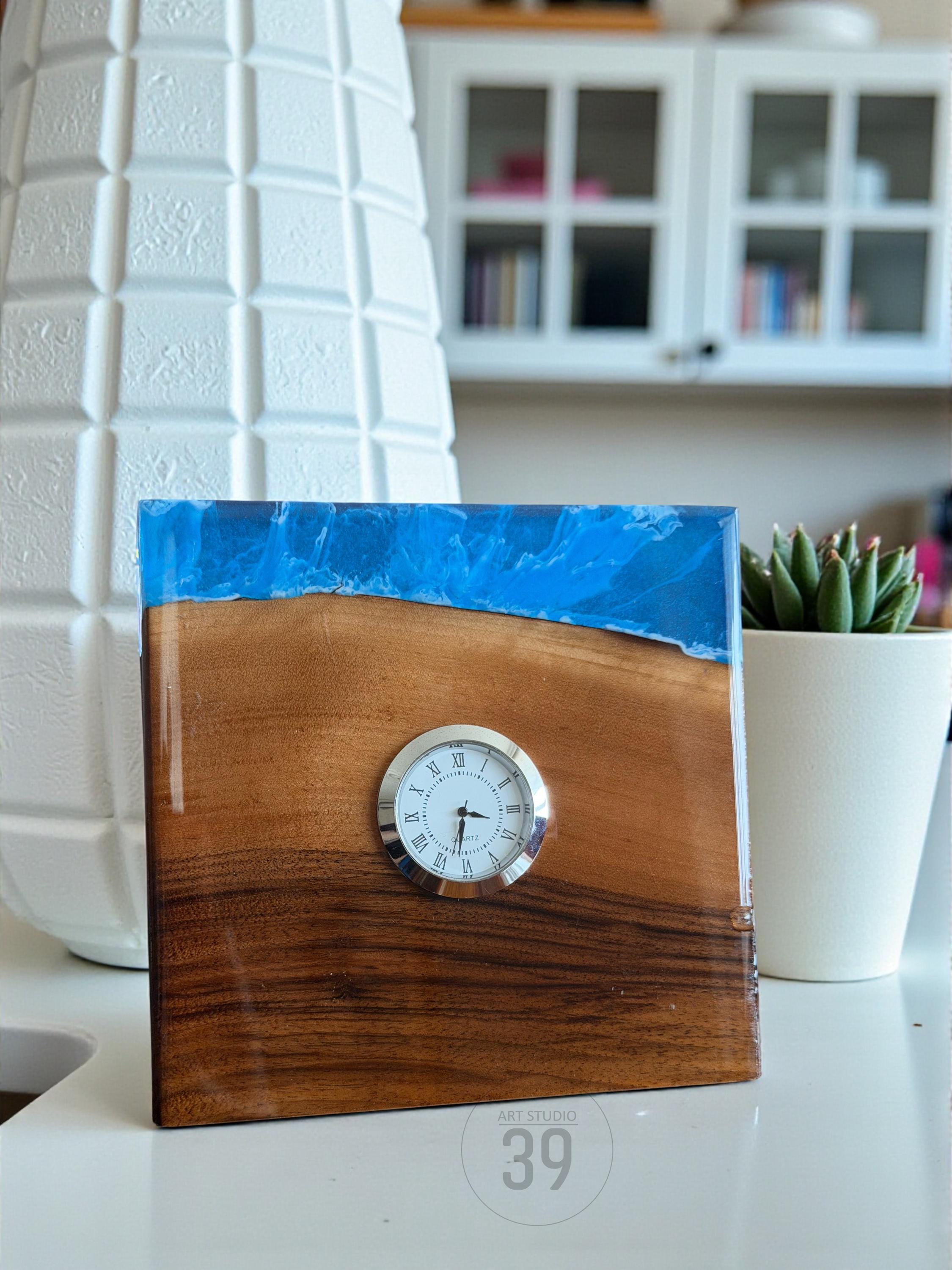 A square wooden clock with a blue resin top. The clock face is white with black Roman numerals and hands. The wood has a rich, brown grain. The clock is labeled "ART STUDIO 39".