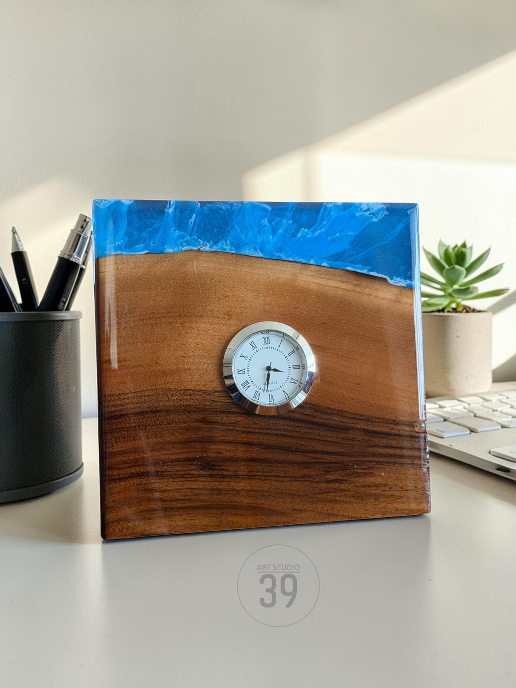 A square wooden desk clock with a blue resin top. The clock face is white with black Roman numerals and hands. The wood is a rich brown color. The clock is labeled "ART STUDIO 39".
