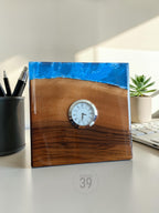 A square wooden desk clock with a blue resin top. The clock face is white with black Roman numerals and hands. The wood is a rich brown color. The clock is labeled "ART STUDIO 39".