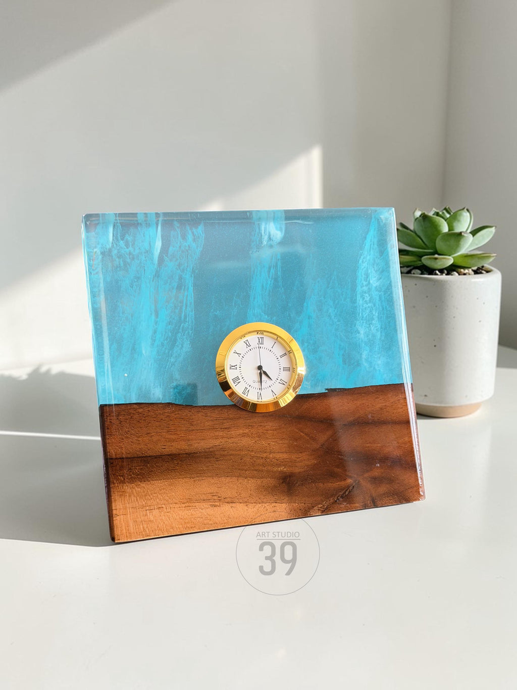 A square desk clock with a gold-colored clock face and Roman numerals. The clock is set in a block of wood and blue resin. The bottom of the clock is wood, and the top is blue resin. The text "ART STUDIO 39" is on the bottom.