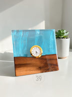 A square desk clock with a gold-colored clock face and Roman numerals. The clock is set in a block of wood and blue resin. The bottom of the clock is wood, and the top is blue resin. The text "ART STUDIO 39" is on the bottom.