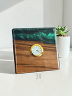 Square wooden clock with a gold-tone clock face and Roman numerals. The top portion is a dark green, swirled resin. The clock is set on a white surface with a small succulent plant in a white pot in the background. The clock has the text "ART STUDIO 39".