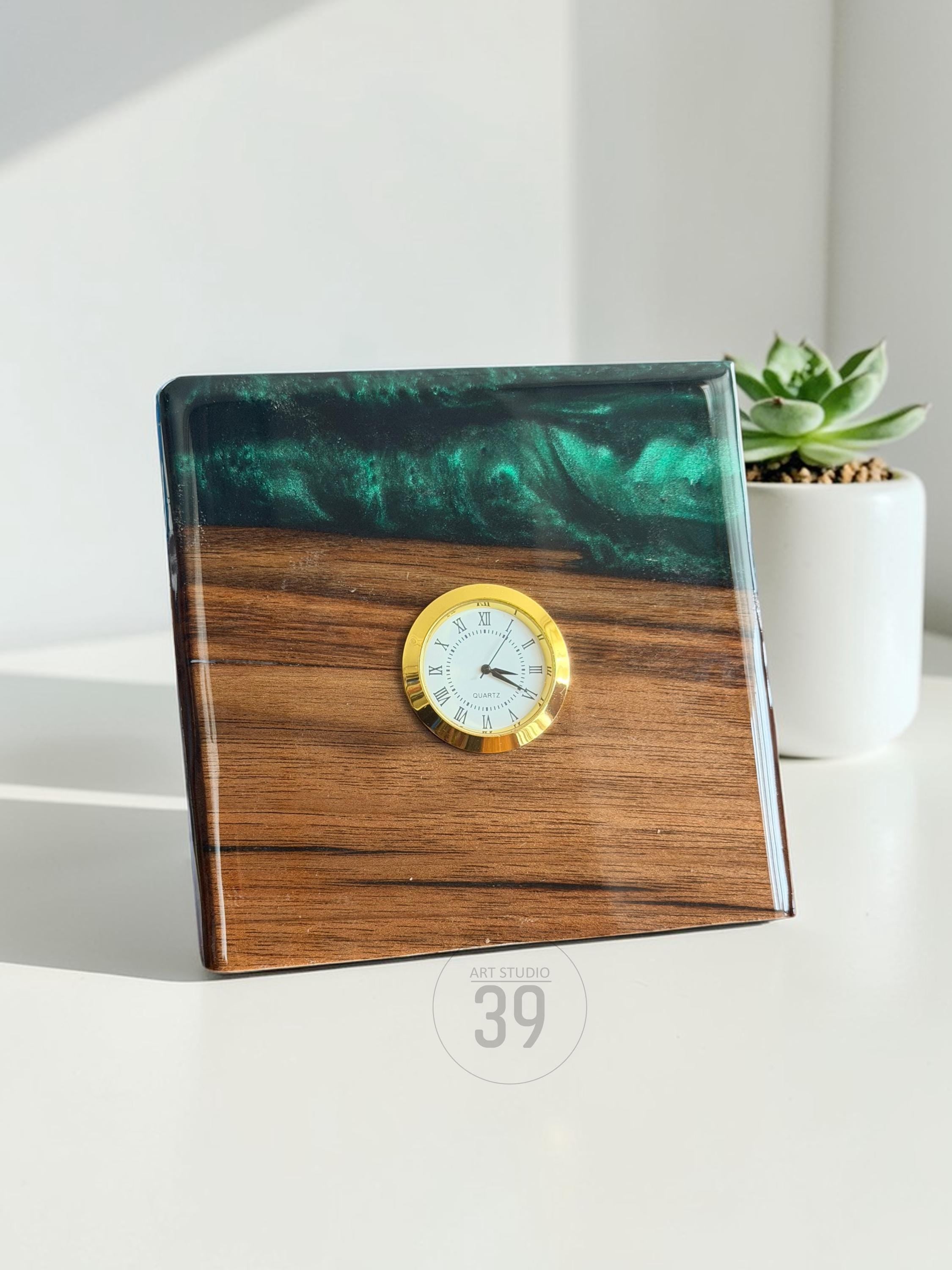 Square wooden clock with a gold-tone clock face and Roman numerals. The top portion is a dark green, swirled resin. The clock is set on a white surface with a small succulent plant in a white pot in the background. The clock has the text "ART STUDIO 39".