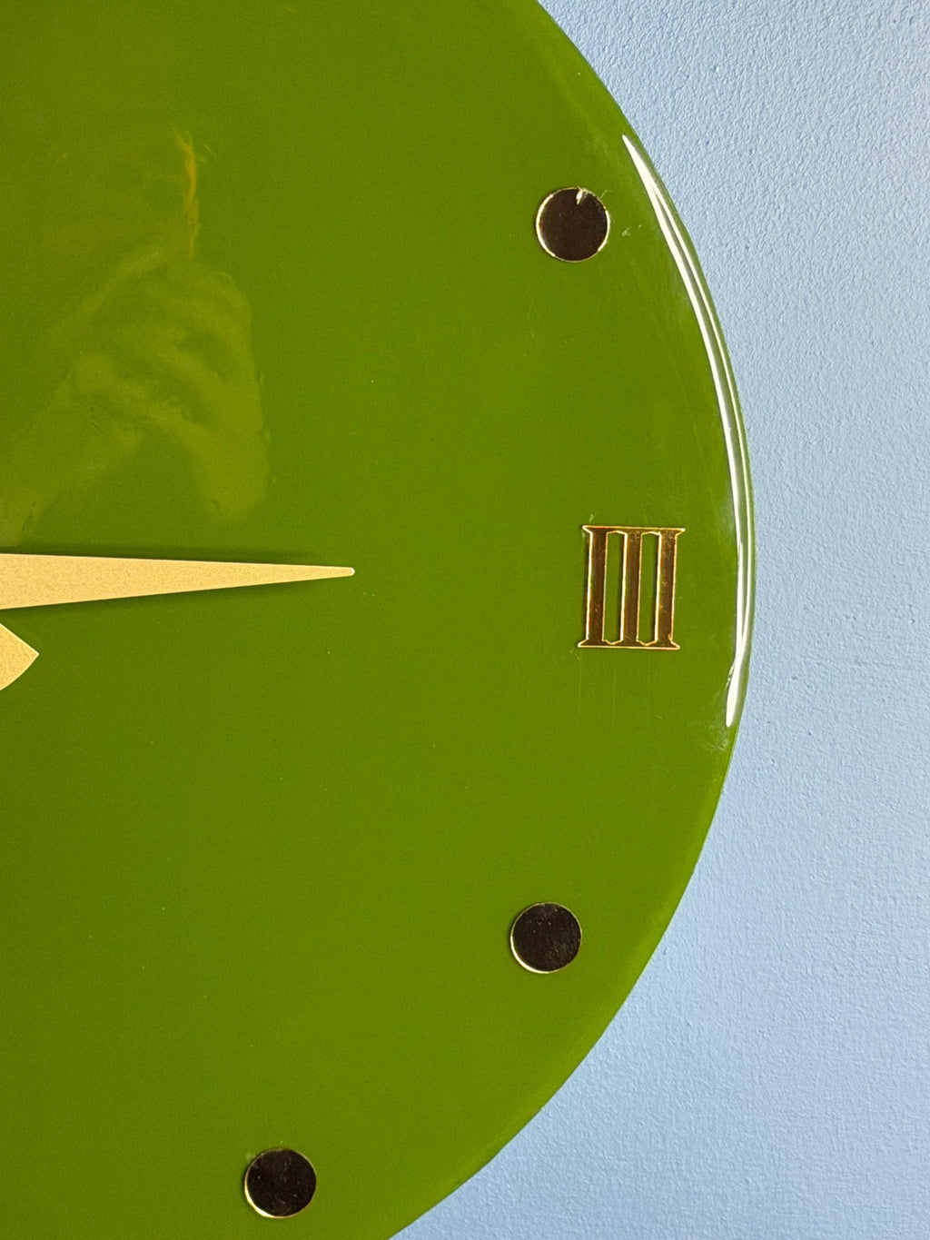 A green wall clock with gold-colored hands and Roman numeral III. The clock face has gold-colored dots marking the hours. The clock is round and has a glossy finish.