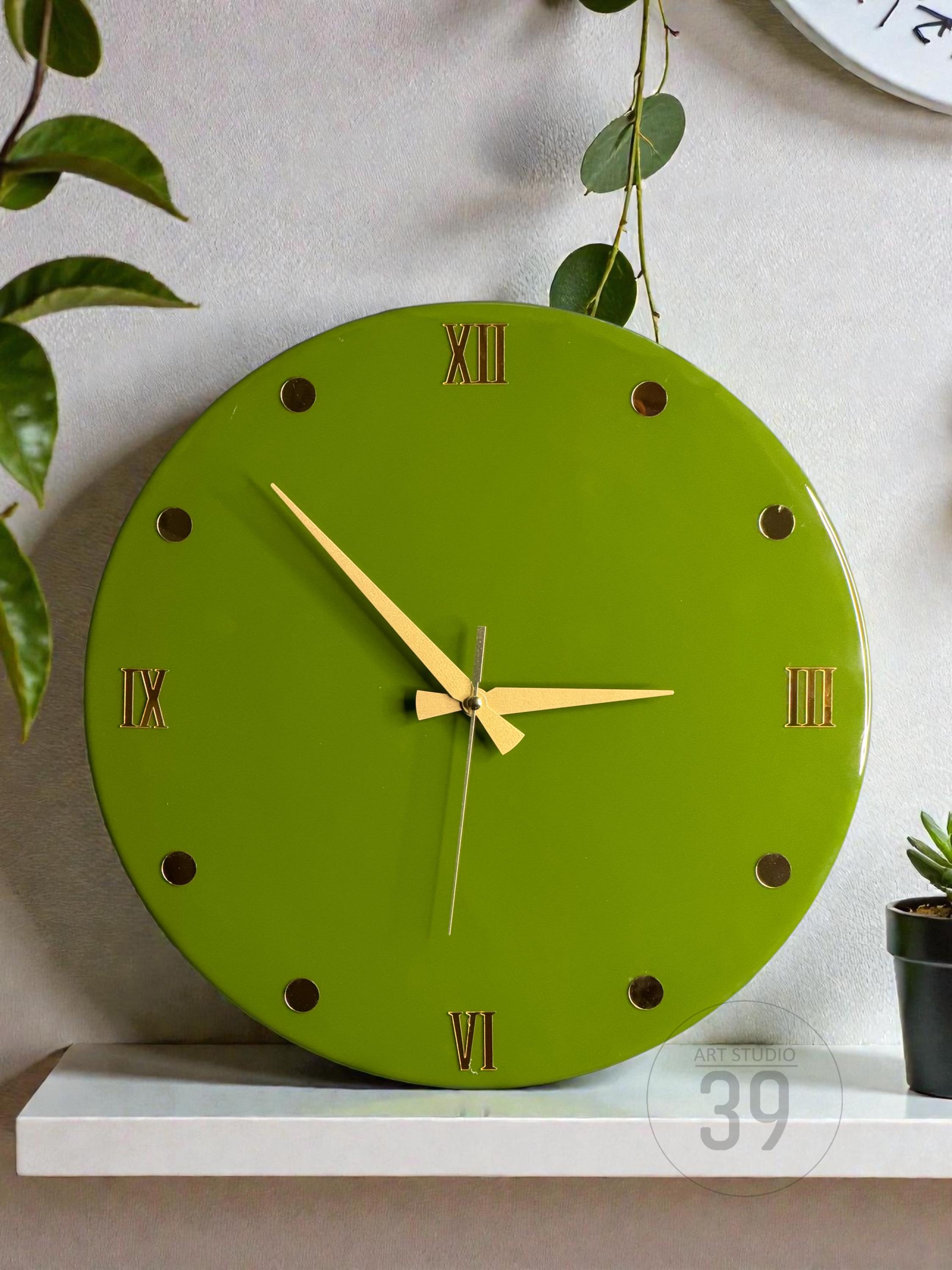 A round, lime green wall clock with gold-colored Roman numerals and hands. The clock has gold-colored dots marking the hours. The clock is on a white shelf with the text "ART STUDIO 39" at the bottom.