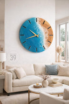 A large, round wall clock with a coastal theme. The clock face features a blue ocean design with a sandy beach and gold-colored hands. The clock is mounted on a white wall in a living room setting. The text "ART STUDIO 39" is visible.