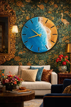 A round wall clock with a beach-themed design. The clock face features a blue ocean and sandy beach, with gold-colored hands and black hour markers. The clock is mounted on a floral wallpaper background.