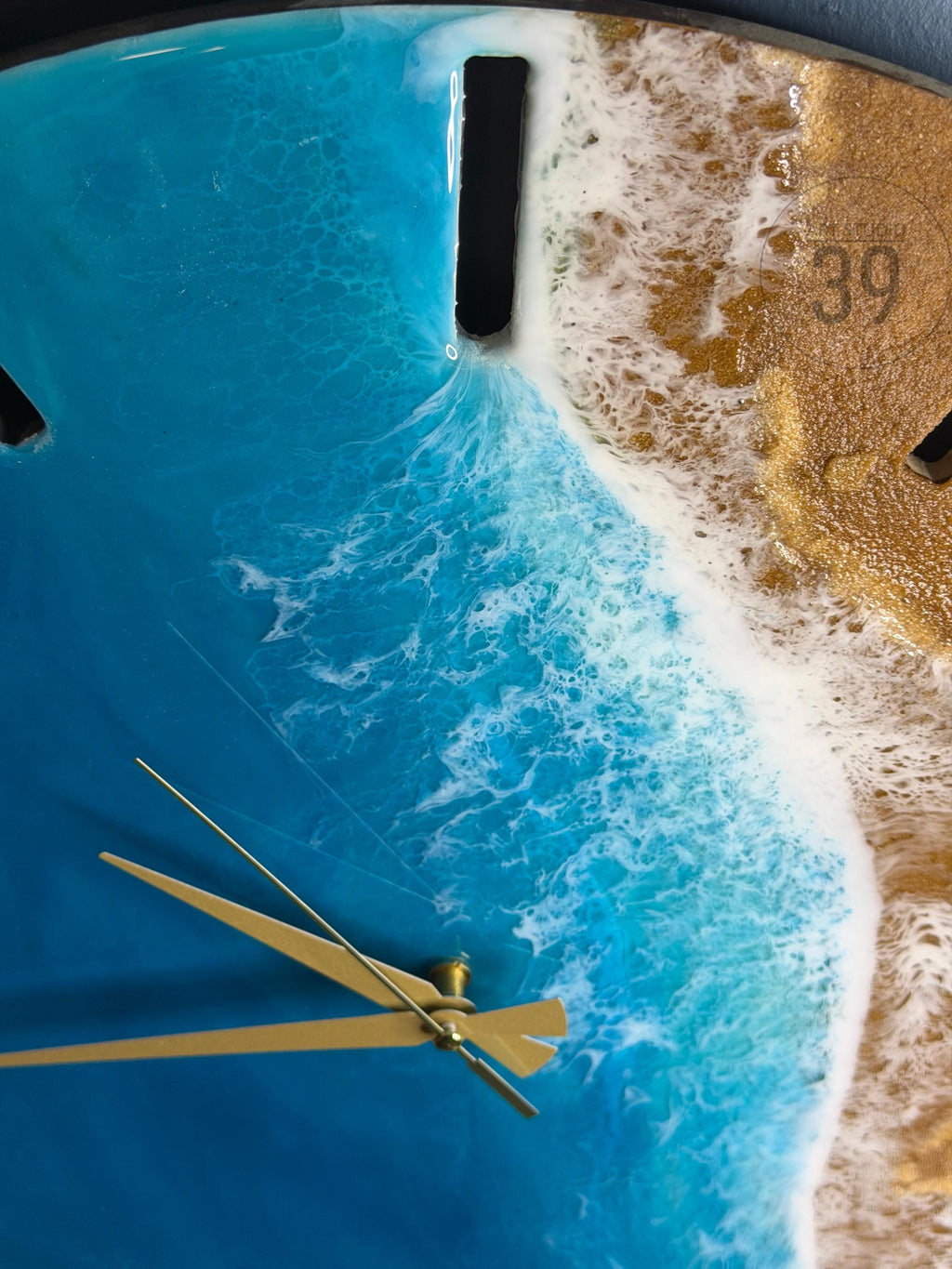A round wall clock with a beach-themed design. The clock face features a blue ocean with white waves and a golden sand-like section. Gold-colored clock hands point to the time. The number 39 is visible on the clock face.