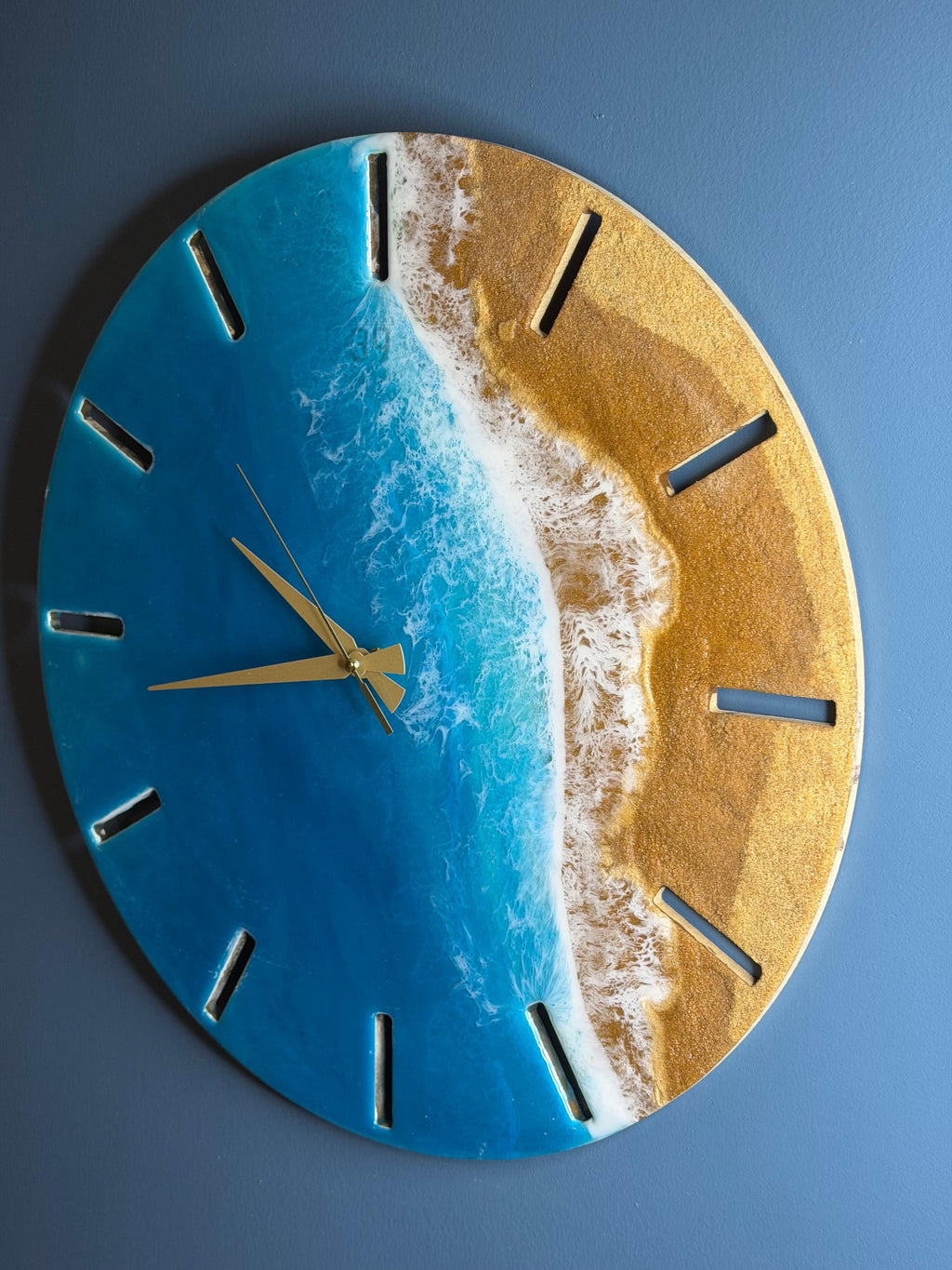 Round wall clock with a beach scene design. The clock face features a blue ocean, white waves, and a golden sand beach. Gold-colored clock hands and hour markers complete the design. The clock is approximately 12 inches in diameter.