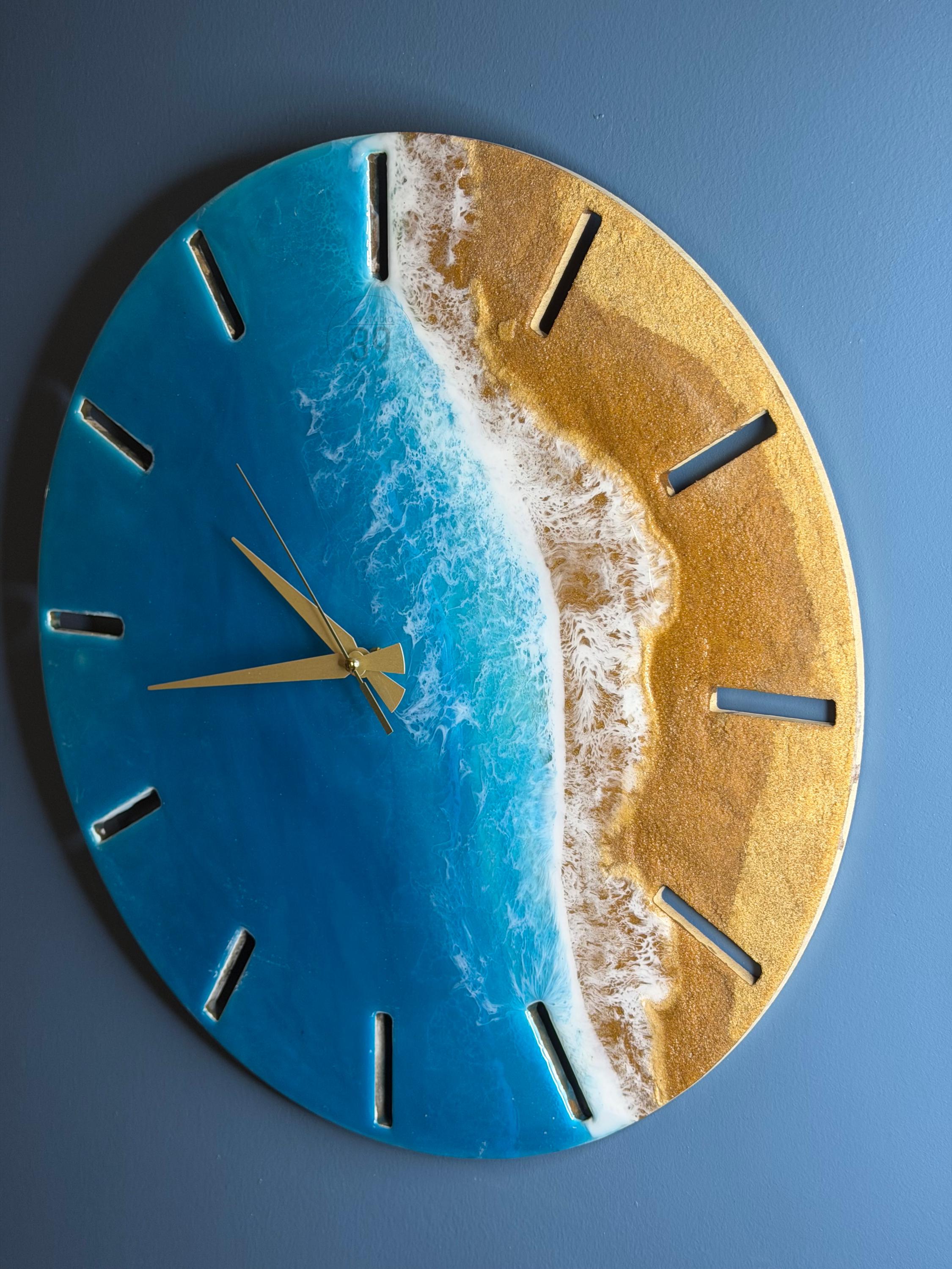 Round wall clock with a beach scene design. The clock face features a blue ocean, white waves, and a golden sand beach. Gold-colored clock hands and hour markers complete the design. The clock is approximately 12 inches in diameter.