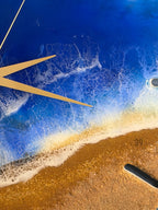 A round clock face with a beach-themed design. The clock features a blue ocean with white foamy waves and a sandy shore. Gold-colored clock hands point to the time. The number 39 is visible on the sand.