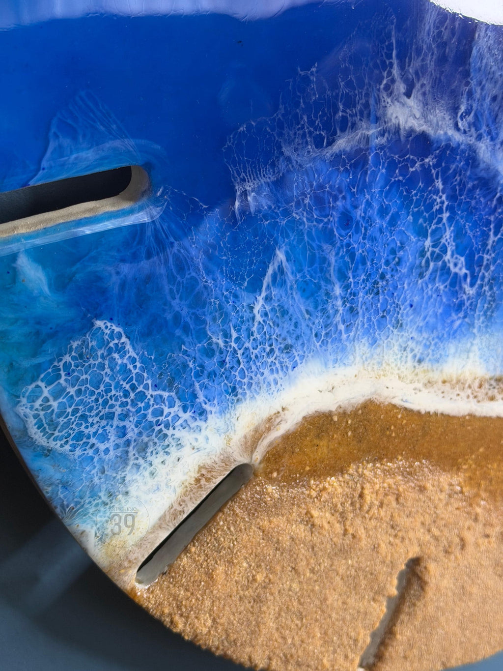 A round, resin art piece depicting a beach scene. The artwork features a deep blue ocean with white foamy waves crashing onto a sandy shore. The piece has two dark, rectangular cutouts and the number "39" is visible.