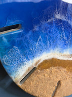 A round, resin art piece depicting a beach scene. The artwork features a deep blue ocean with white foamy waves crashing onto a sandy shore. The piece has two dark, rectangular cutouts and the number "39" is visible.