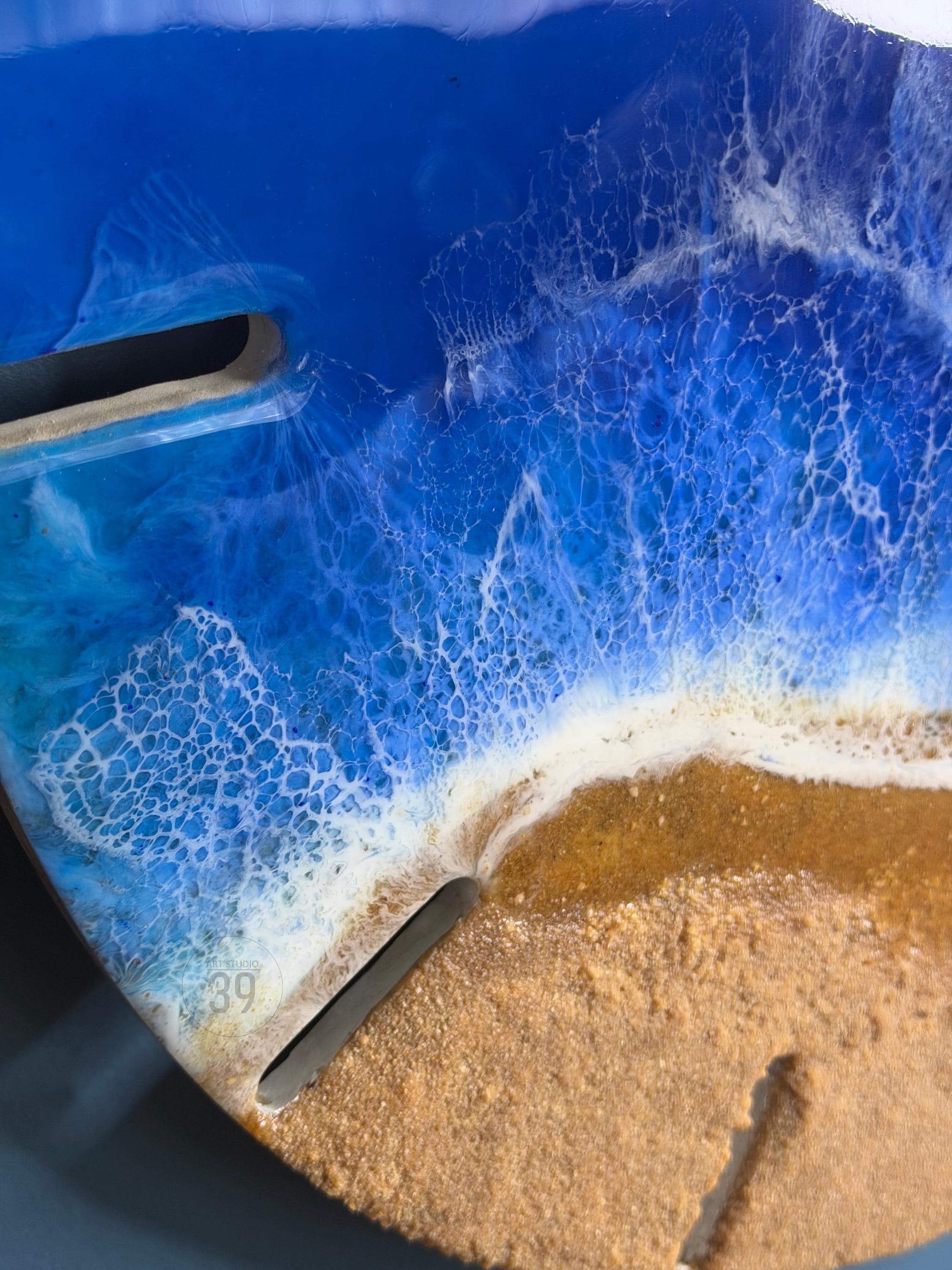 A round, resin art piece depicting a beach scene. The artwork features a deep blue ocean with white foamy waves crashing onto a sandy shore. The piece has two dark, rectangular cutouts and the number "39" is visible.