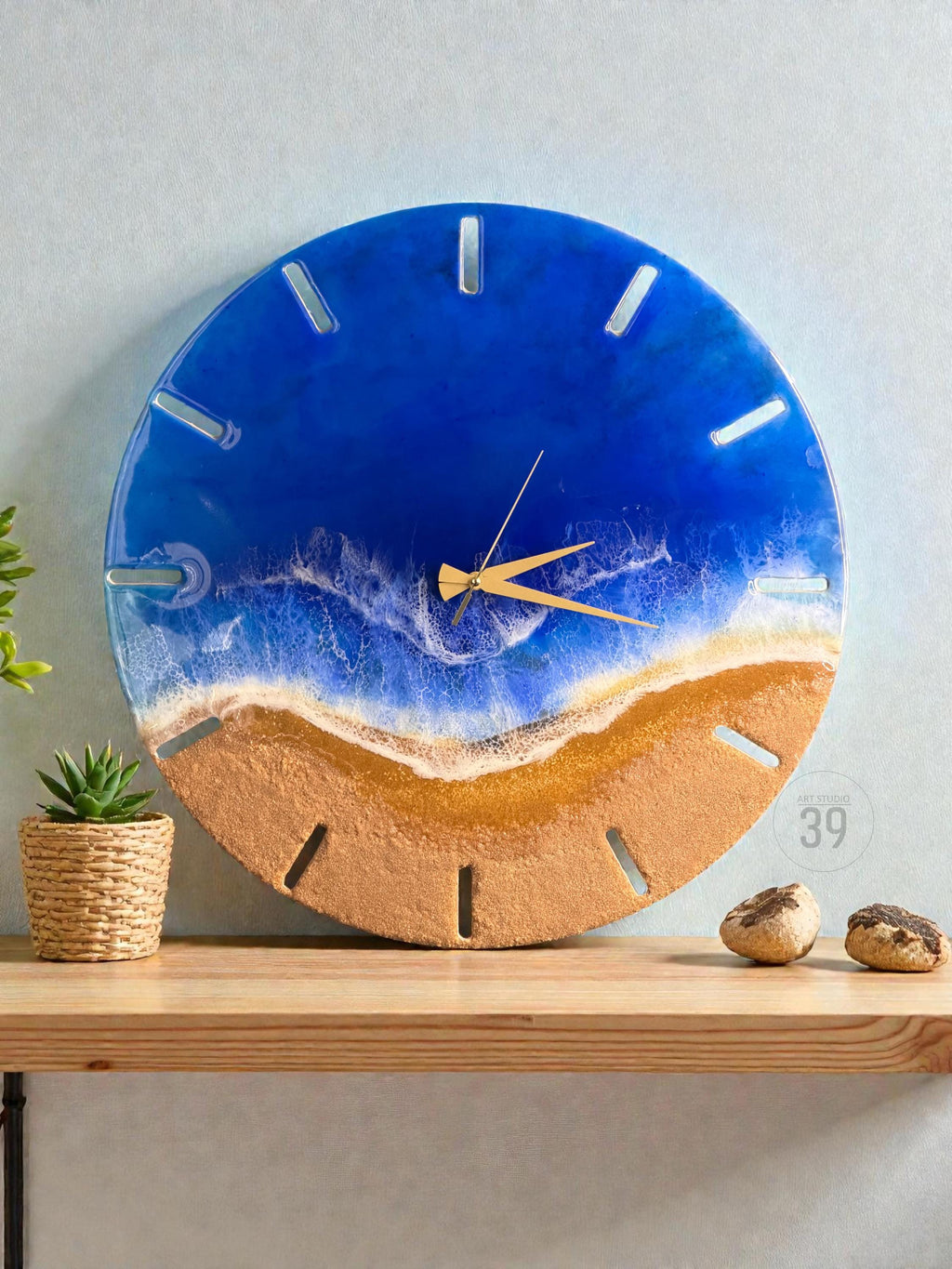 A round wall clock with a beach-themed design. The top half is a deep blue, resembling the ocean, while the bottom half is a sandy brown. The clock hands are gold, and the hour markers are rectangular cutouts.