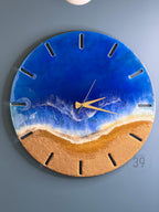 Round wall clock with a beach-themed design. The clock face features a blue ocean, white foamy waves, and a golden sand-colored beach. Gold-colored clock hands and hour markers complete the design. The clock is mounted on a blue wall.