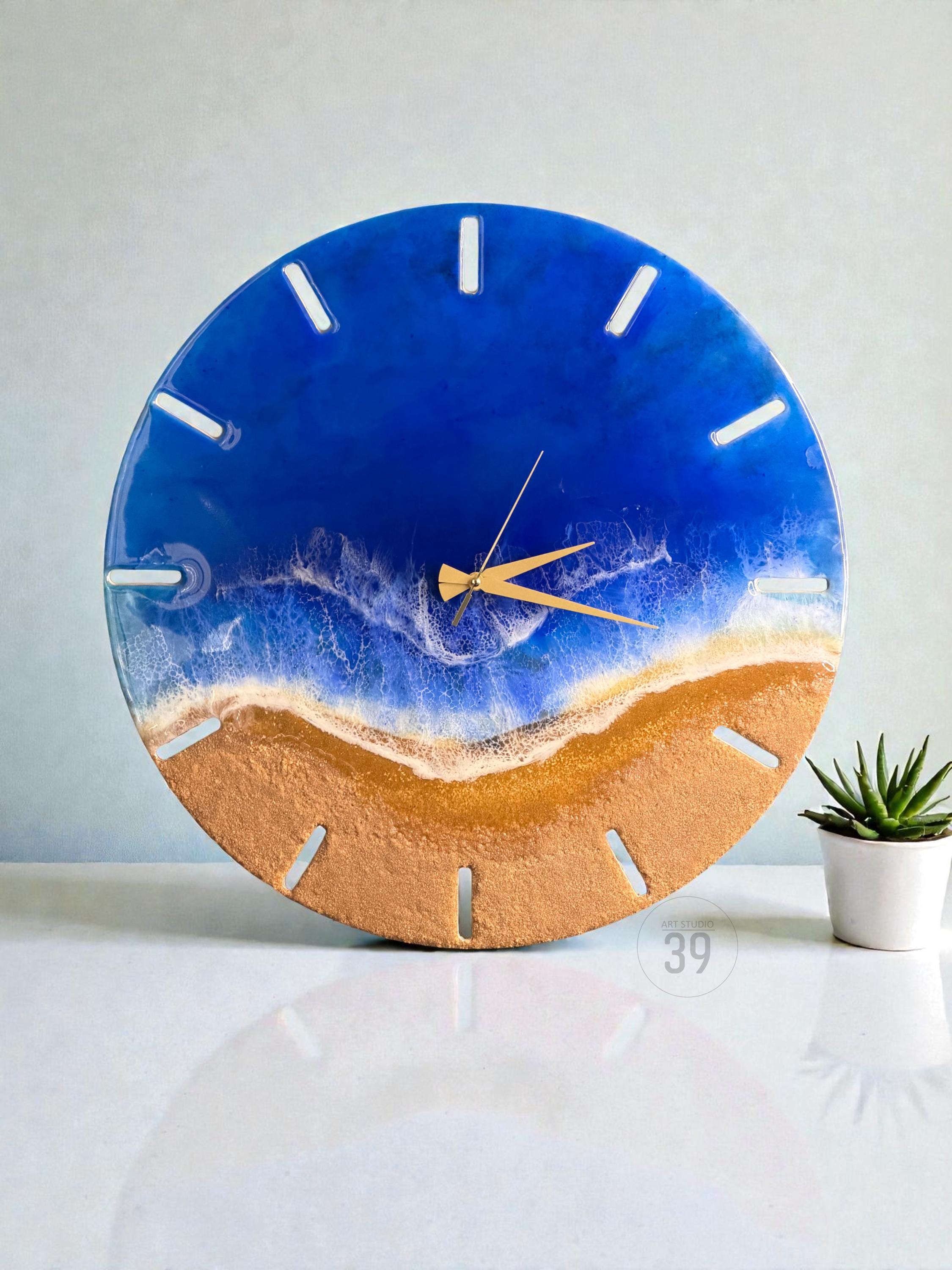 A round wall clock with a beach-themed design. The top half is a deep blue, resembling the ocean, while the bottom half is a sandy gold color. The clock hands are gold, and the hour markers are white. A small succulent plant sits to the right.