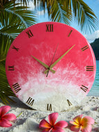 A round wall clock with a pink and white resin design, featuring gold Roman numerals and hands. The clock has a beach-themed aesthetic, with a gradient effect resembling sand and water. The clock is approximately 12 inches in diameter.