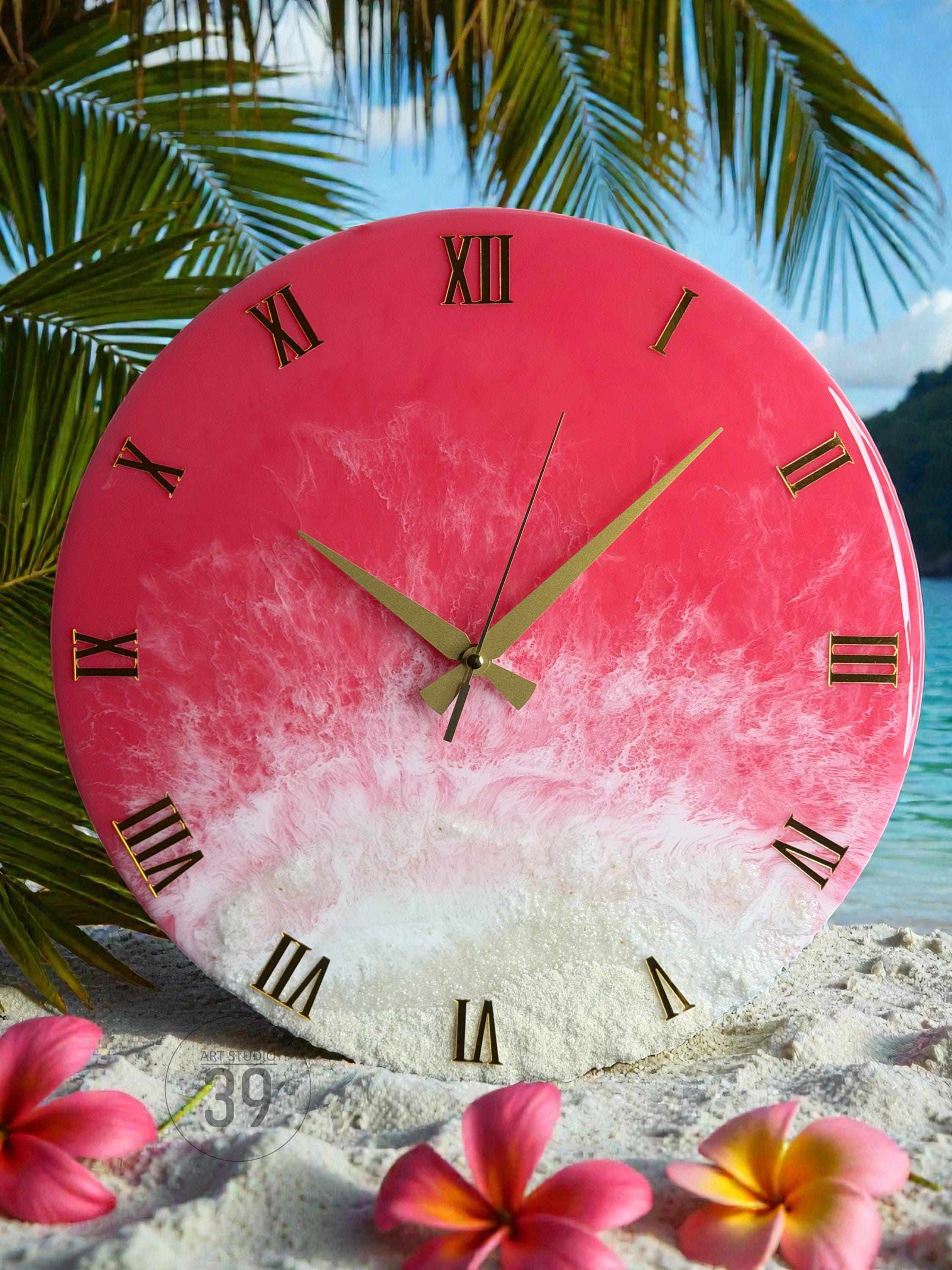 A round wall clock with a pink and white resin design, featuring gold Roman numerals and hands. The clock has a beach-themed aesthetic, with a gradient effect resembling sand and water. The clock is approximately 12 inches in diameter.