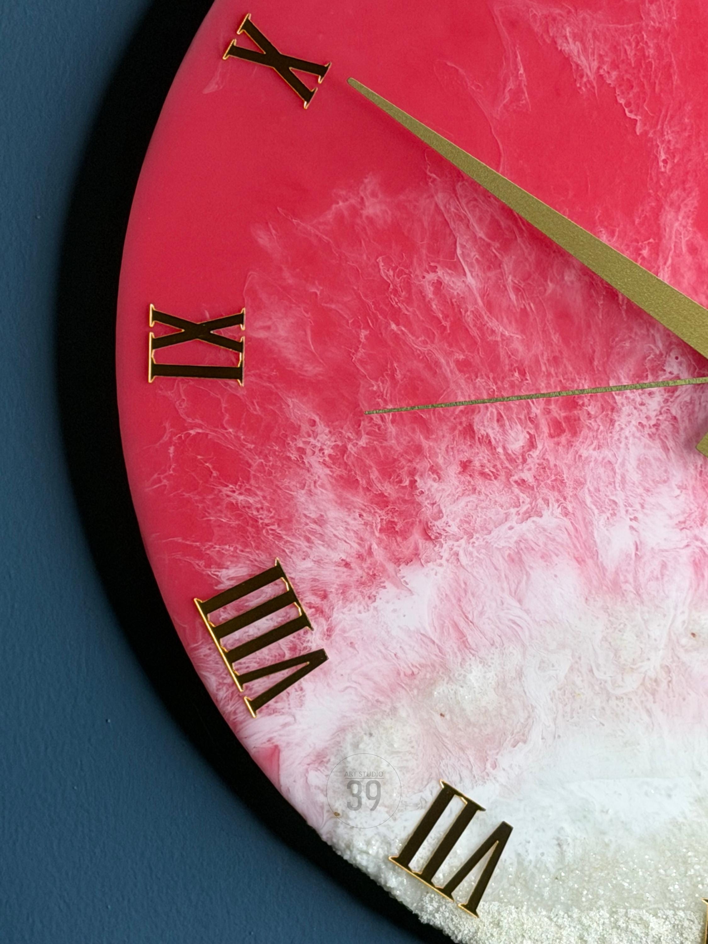 A close-up of a round wall clock with a pink and white marbled design. The clock features gold-colored Roman numerals and hands. The clock's face has a textured, ocean-like appearance.
