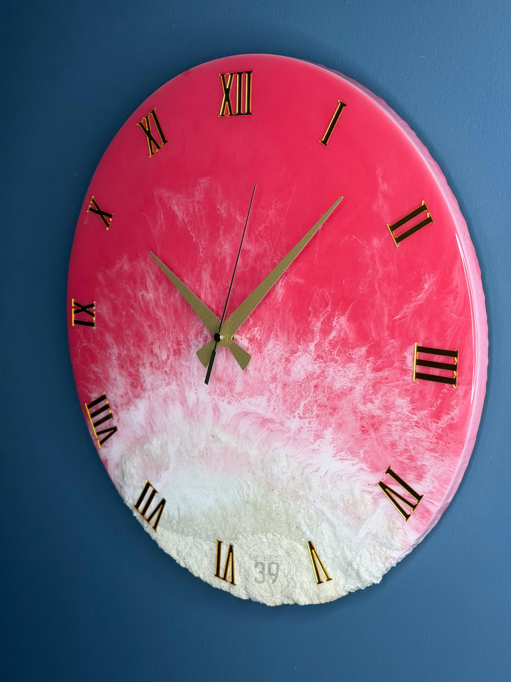 A round wall clock with a pink and white resin design. The clock features gold Roman numerals and hands. The clock's face has a textured, wave-like appearance, with the number 39 visible near the bottom.