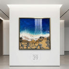 A framed artwork depicting an aerial view of a beach scene. The artwork features a deep blue ocean, white foamy waves, a sandy shore, and rocky areas. The frame is gold-colored, and the artwork is displayed on a white wall.