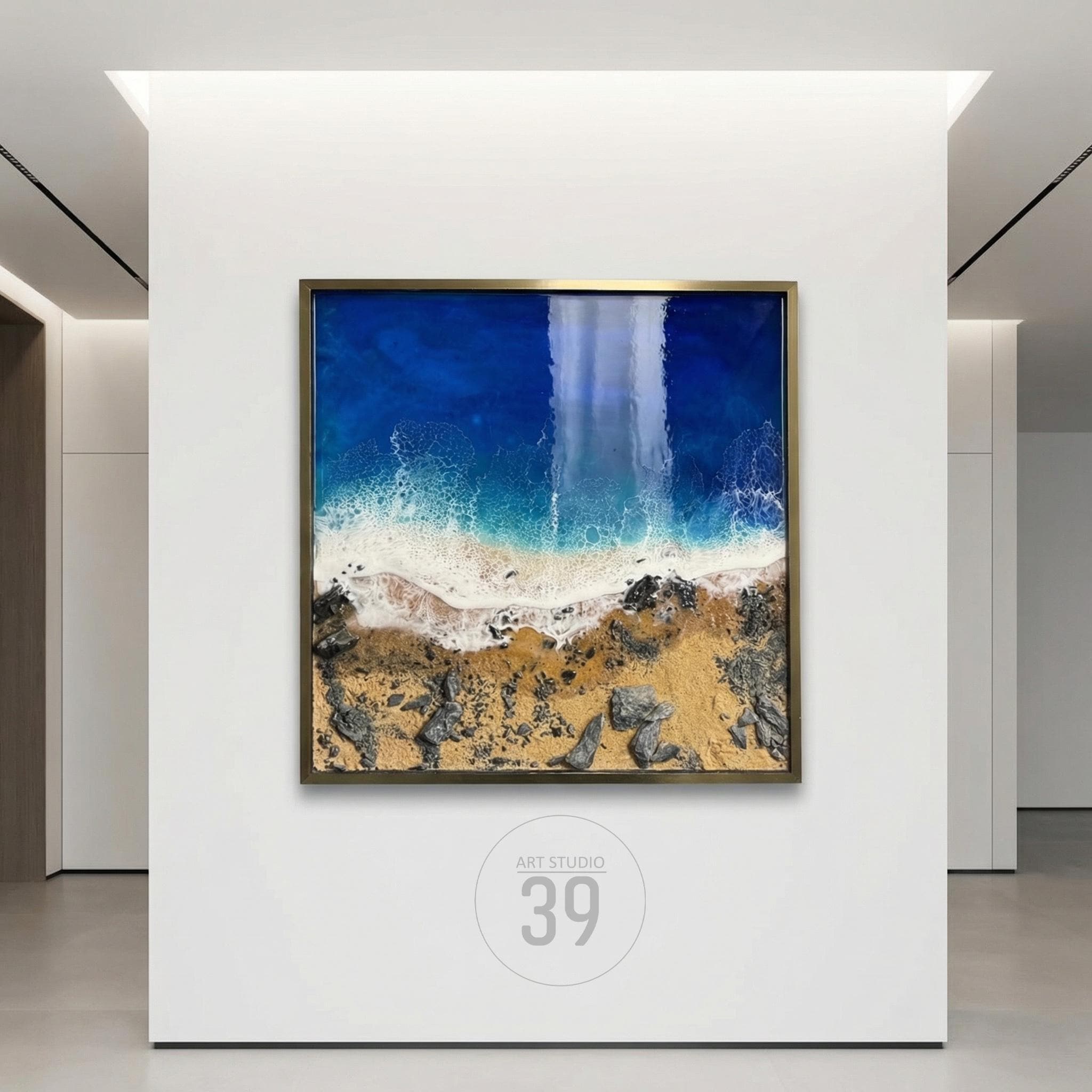 A framed artwork depicting an aerial view of a beach scene. The artwork features a deep blue ocean, white foamy waves, a sandy shore, and rocky areas. The frame is gold-colored, and the artwork is displayed on a white wall.