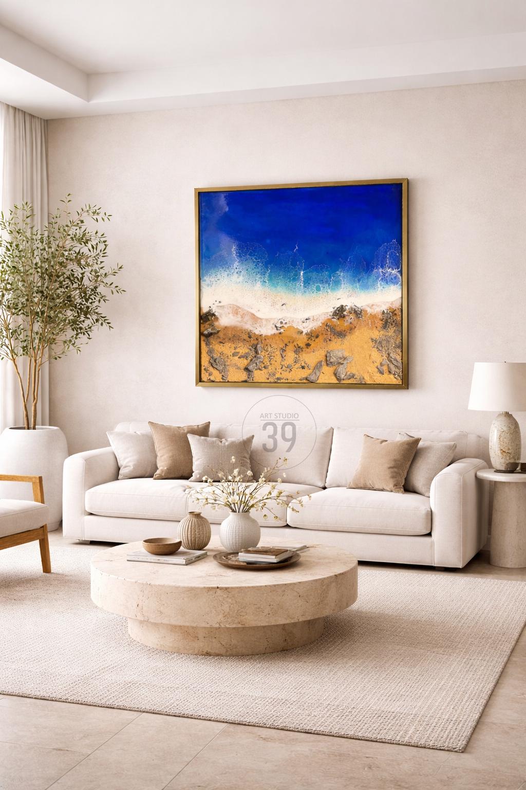 A modern living room with a large framed abstract painting of a beach scene. The painting features a blue ocean, white waves, and a sandy shore. The room includes a white sofa, coffee table, and decorative pillows.