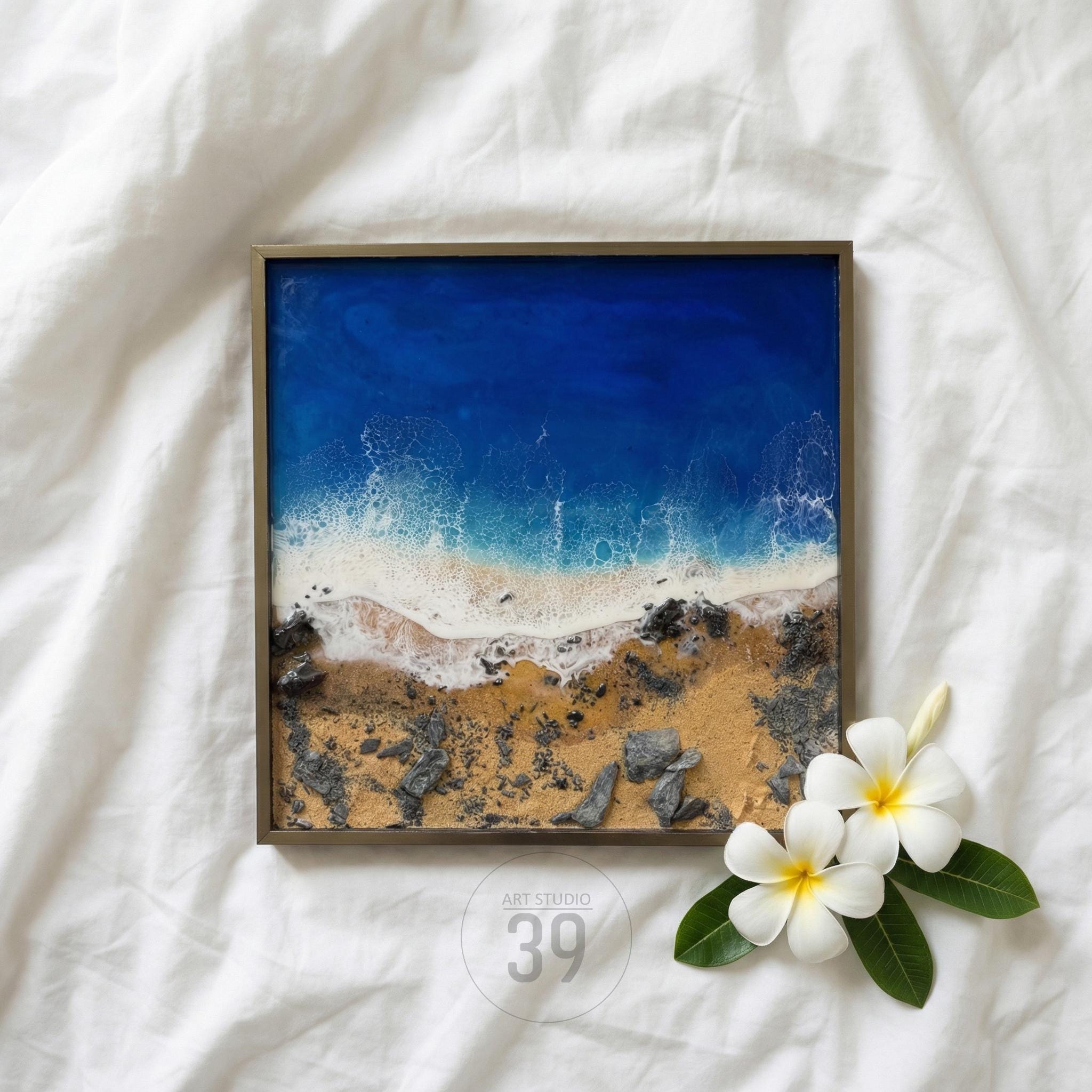 Square framed artwork depicting a beach scene with a deep blue ocean, white foamy waves, and a sandy shore with rocks. The artwork is accented with white flowers and green leaves. The artwork is labeled "ART STUDIO 39".