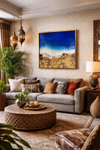 Living room with a gray sofa, decorative pillows, and a round wooden coffee table. A framed abstract painting of a beach scene hangs above the sofa. Decorative lanterns and a patterned rug complete the space.