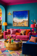 A vibrant living room with a pink sofa, blue armchair, and a gold coffee table. The walls are teal, and the ceiling is magenta. A framed artwork depicts a beach scene. Decorative pillows and lamps add color.