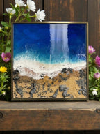Square framed resin art depicting a beach scene. The artwork features a deep blue ocean, white foamy waves, sandy shore, and dark rocks. The frame is a muted gold color. Flowers surround the artwork.