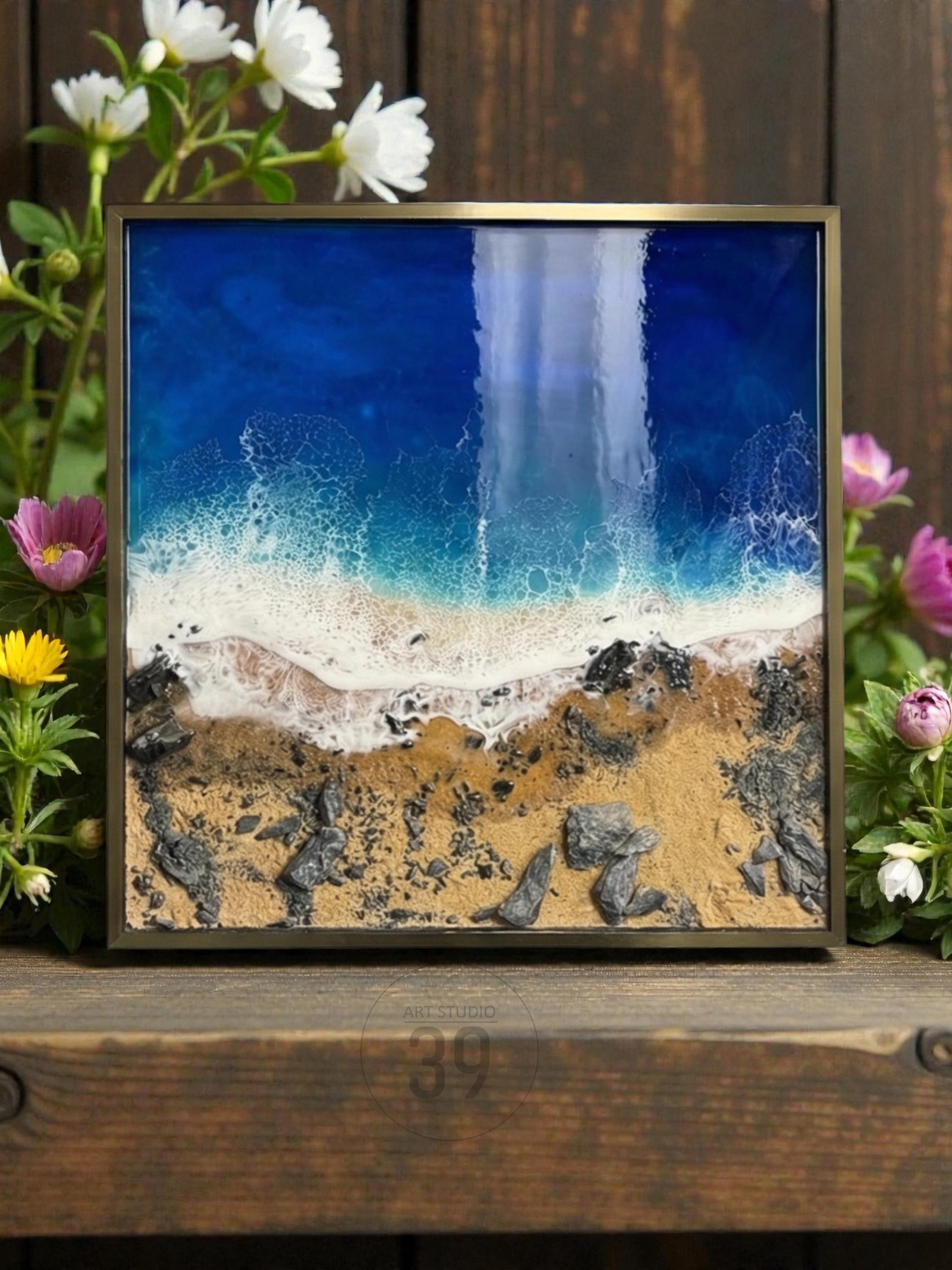 Square framed resin art depicting a beach scene. The artwork features a deep blue ocean, white foamy waves, sandy shore, and dark rocks. The frame is a muted gold color. Flowers surround the artwork.