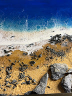 A resin art piece depicting a beach scene. The artwork features a deep blue ocean, foamy white waves, and a sandy shore with scattered dark gray rocks. The piece has a textured surface and a glossy finish.