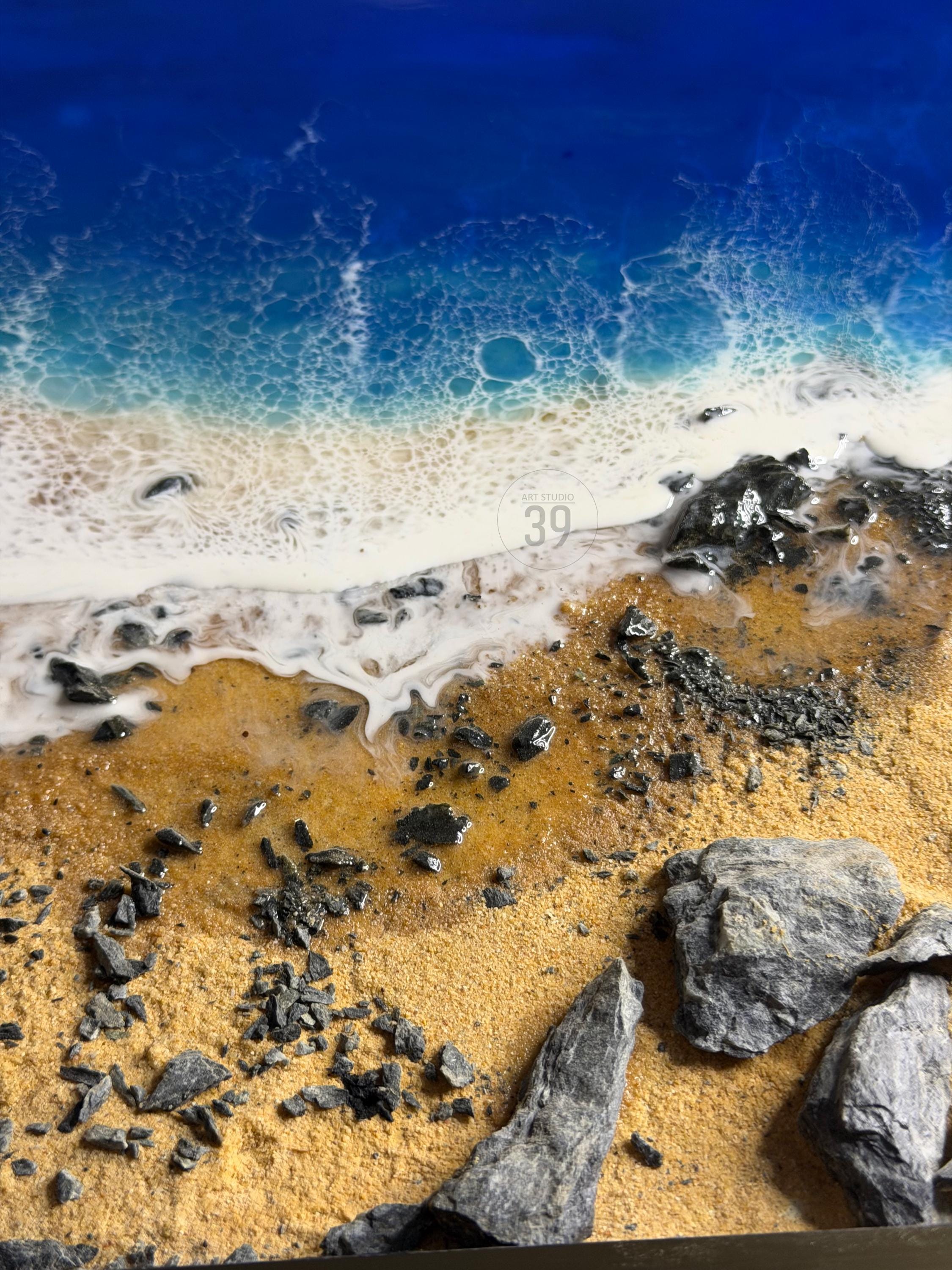 A resin art piece depicting a beach scene. The artwork features a deep blue ocean, foamy white waves, and a sandy shore with scattered dark gray rocks. The piece has a textured surface and a glossy finish.