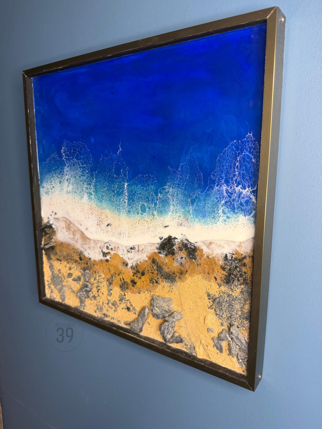 Abstract framed artwork depicting a beach scene. The top portion shows a deep blue ocean, transitioning to white foamy waves. The bottom features a sandy beach with rocks, all within a dark metal frame.