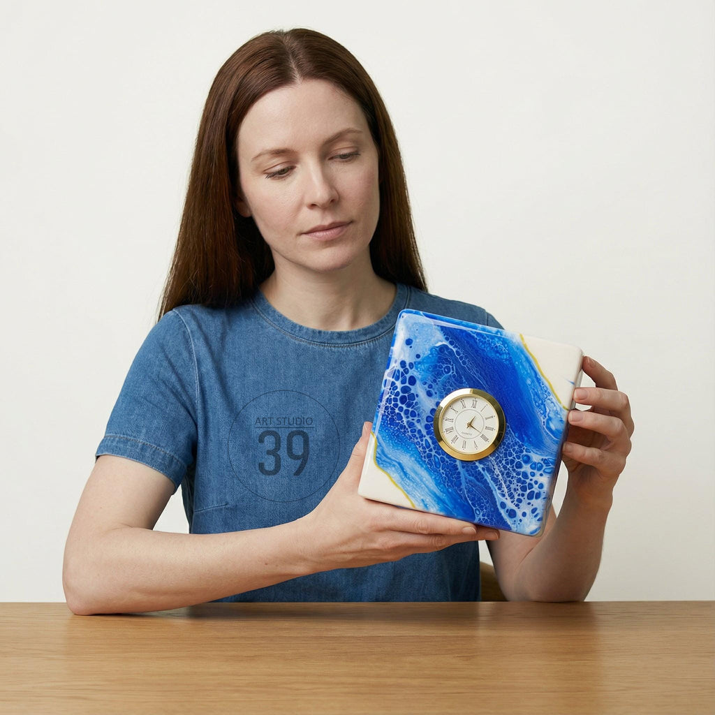 Square clock with a white face and gold-tone trim, set within a blue and white abstract design. The clock is held by a person wearing a blue denim shirt. The clock's face displays Roman numerals.