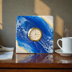 A square clock with a gold-colored bezel and a cream-colored face. The clock is set into a block with a blue and white abstract design. A white coffee cup and saucer sit to the right of the clock on a wooden surface.