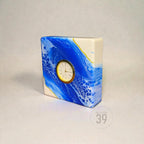 A rectangular clock with a white face and gold-colored trim, set within a blue and white abstract design. The clock's face features Roman numerals. The base is white with blue and gold accents. The clock is labeled "ART STUDIO 39".