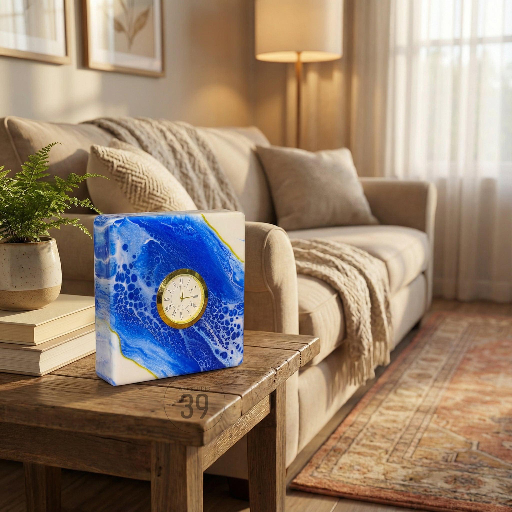 A square clock with a blue and white abstract design, accented with gold. The clock face is gold with black hands and numerals. The clock is sitting on a wooden table.