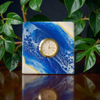 Square clock with a gold-tone bezel and a cream-colored face, set against a blue and white abstract design. The clock is reflected on a dark surface, with green leaves in the background. The clock face features Roman numerals.