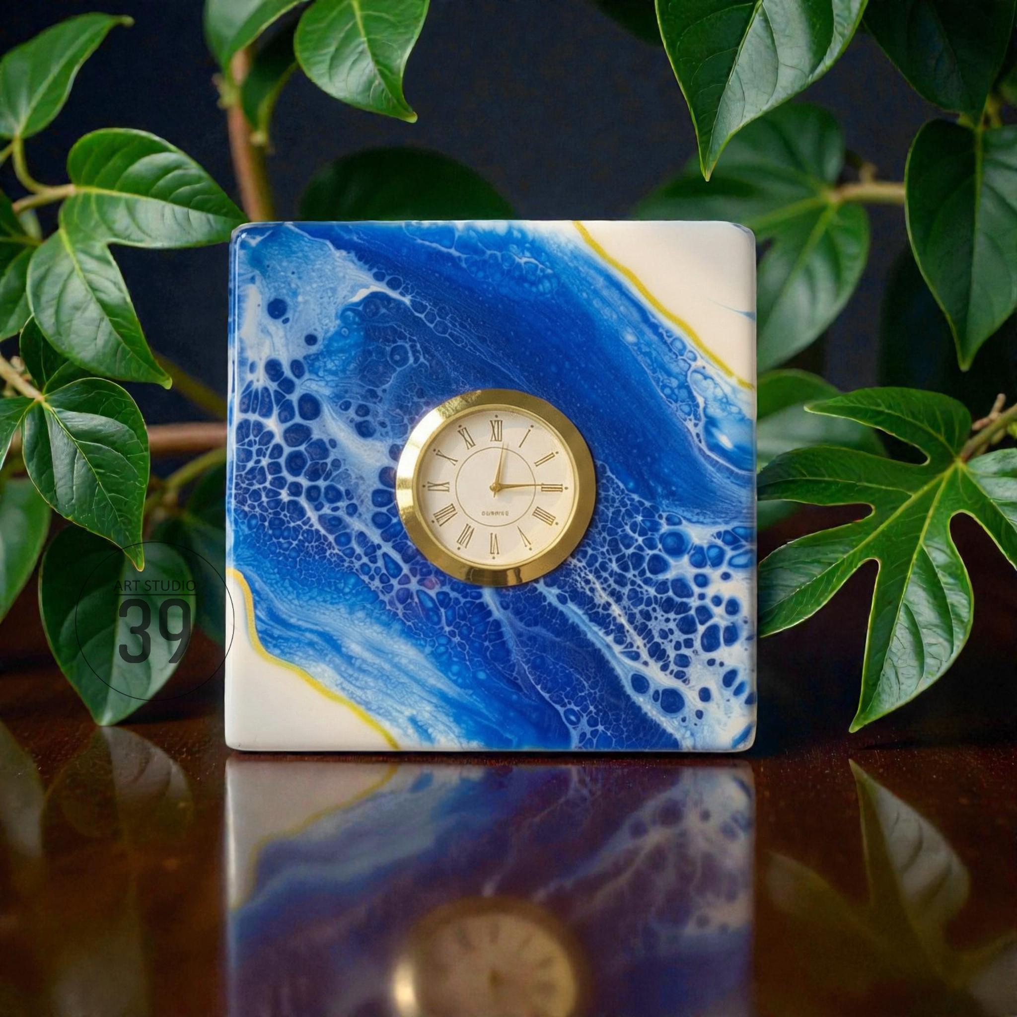 Square clock with a gold-tone bezel and a cream-colored face, set against a blue and white abstract design. The clock is reflected on a dark surface, with green leaves in the background. The clock face features Roman numerals.