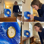 A square clock with a gold-tone bezel and a white face with black Roman numerals. The clock is set into a white and blue abstract painted design. The clock is displayed on a wooden surface.